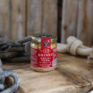 A 185g tin of Shines Chilli Tuna in Oil rests on a rustic wooden surface, surrounded by thick ropes and chains against a backdrop of wooden planks.