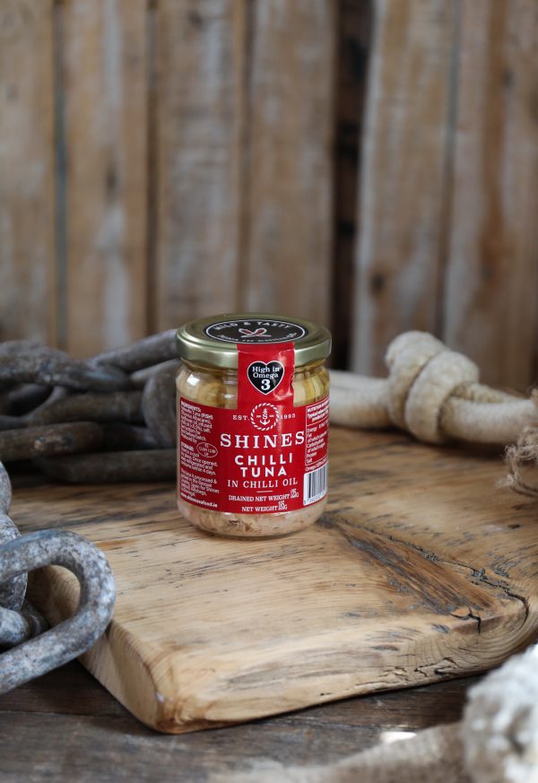 A 185g tin of Shines Chilli Tuna in Oil rests on a rustic wooden surface, surrounded by thick ropes and chains against a backdrop of wooden planks.