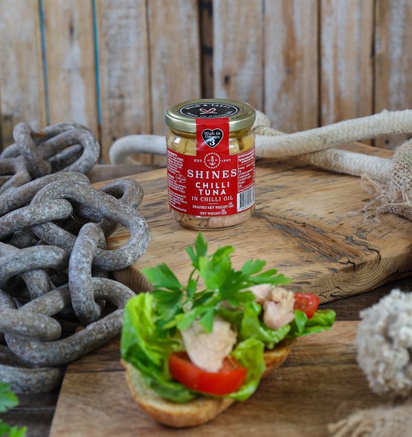 A jar of Shines Chilli Tuna in Oil - 185g rests on a wooden board with rustic chains and rope, while an open sandwich topped with lettuce, tomato, parsley, and chilli tuna is artfully presented in the foreground.