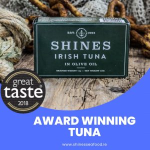 A 111g tin of Shines Wild Irish Tuna in Olive Oil sits on a rustic wooden surface with fishing nets and ropes, featuring a "Great Taste 2018" badge, "Award Winning Tuna," and www.shinesseafood.ie.