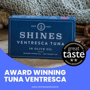 A blue 111g tin of Shines Tuna Ventresca in Olive Oil sits on a wooden surface. Featuring the "Great Taste 2022" award badge, it highlights its status as "Award-Winning Tuna Ventresca." Visit shinessafood.ie for more info.
