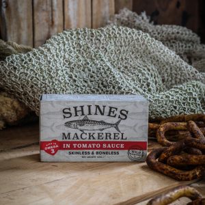 A box of Mackerel Fillets in Tomato Sauce 125g sits on a wooden surface beside fishing nets and rusty metal rings, set against a rustic wooden background.
