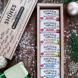 A rectangular "Mackerel Gift Box" holds neatly arranged tins of mackerel, surrounded by festive decorations, pine branches, a wrapped present, baubles, and scattered artificial snow on a wooden surface.