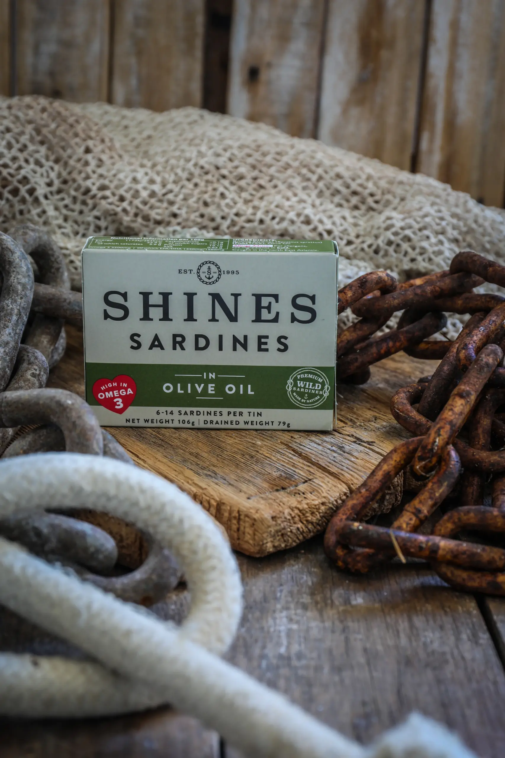 A box of Shines Wild Sardines in Sunflower Oil (106g) rests on a rustic wooden surface, surrounded by thick ropes, rusty chains, and fishing nets for a nautical or fishing-themed setting.