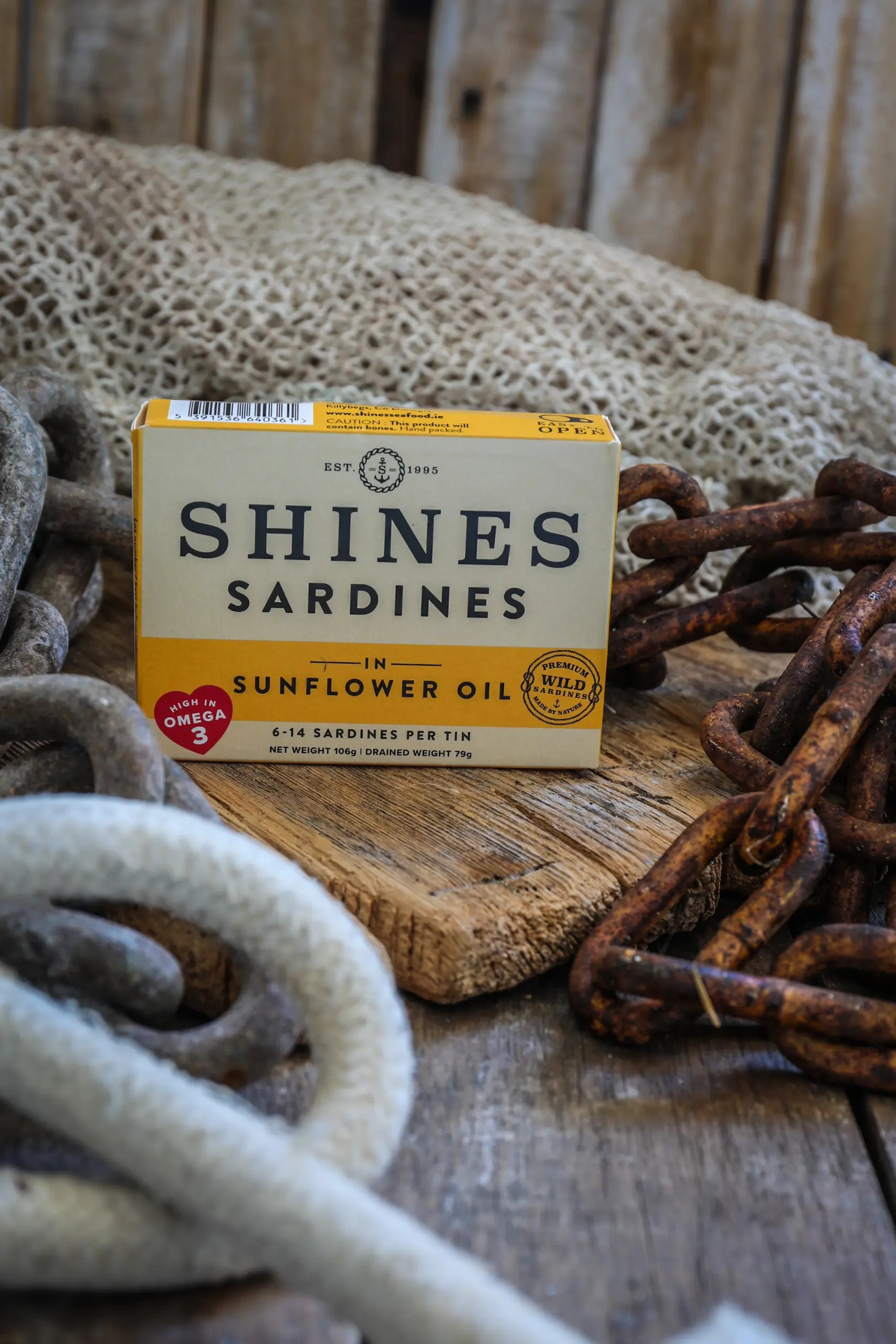 A box of Shines Wild Sardines in Sunflower Oil - 106g rests on a wooden surface, surrounded by ropes, chains, and fishing netting for a rustic nautical atmosphere.