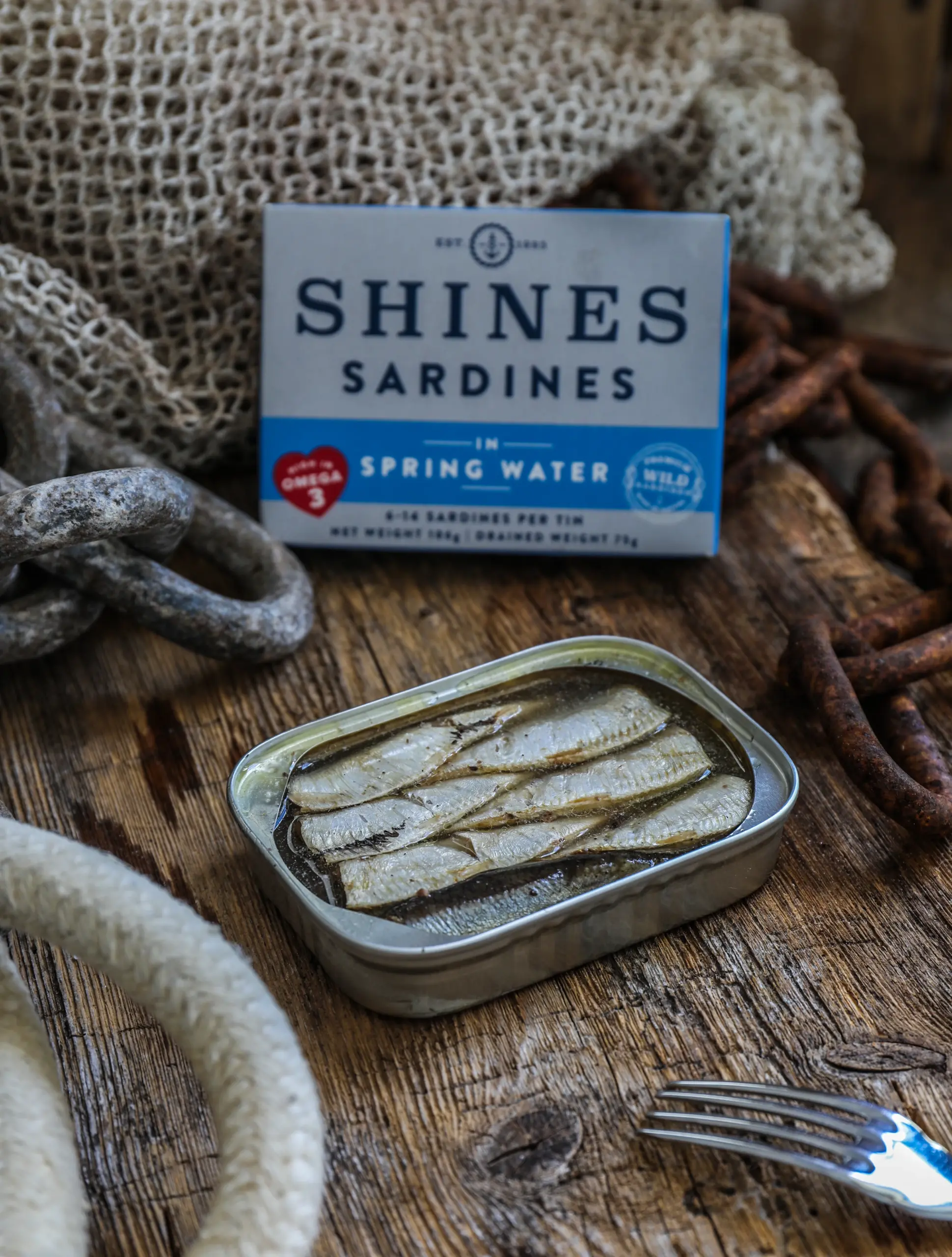 An open tin of Shines Wild Sardines in Sunflower Oil (106g) rests on a rustic wooden surface, with a "Shines Sardines" box and nautical ropes in the background and a fork placed nearby.