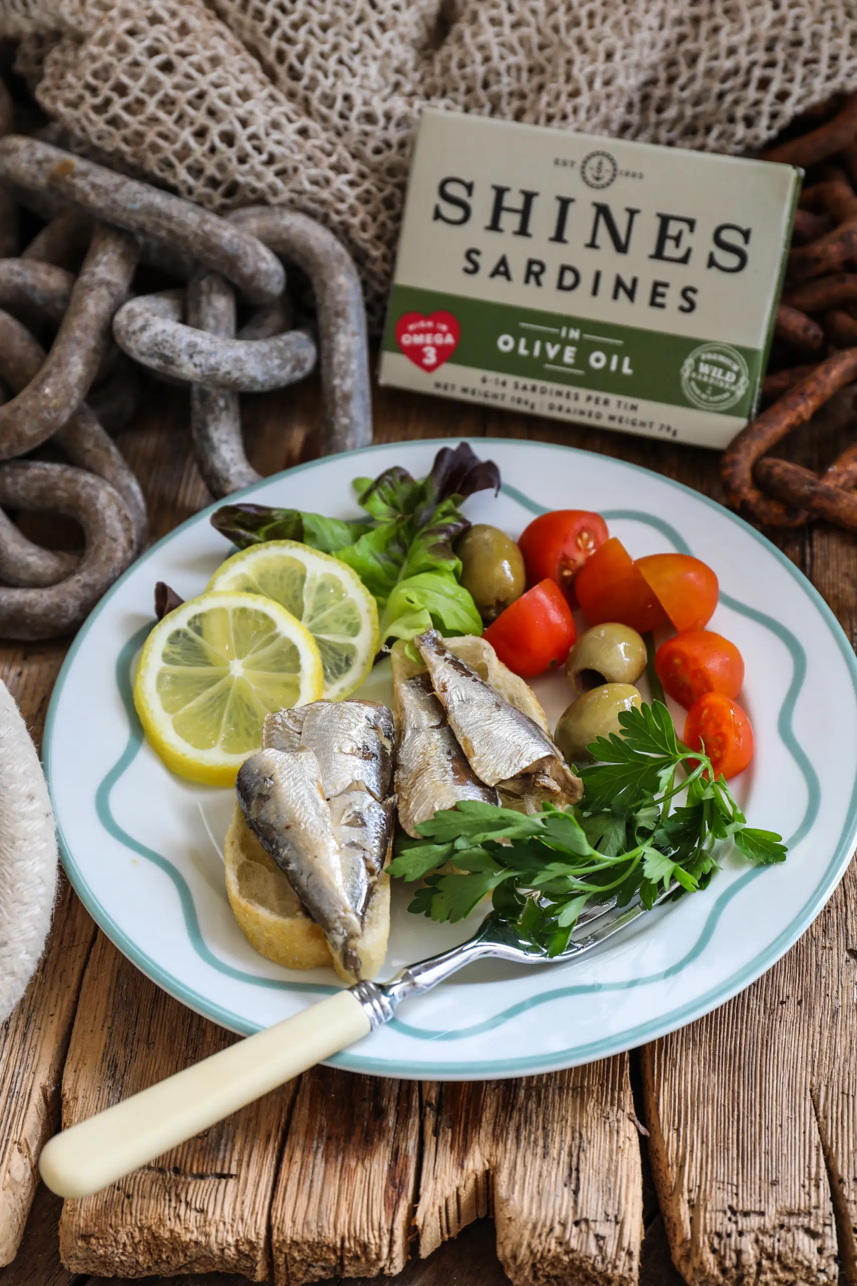A plate with Shines Wild Sardines in sunflower oil on bread, lemon slices, mixed leaves, olives, and cherry tomatoes. A fork rests on the plate; rustic decor and a box of Shines Wild Sardines are in the background.