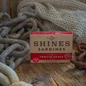 A box of Shines Wild Sardines in Tomato Sauce - 106g rests on a rustic wooden surface, surrounded by thick metal chains, rope, and a fishing net.