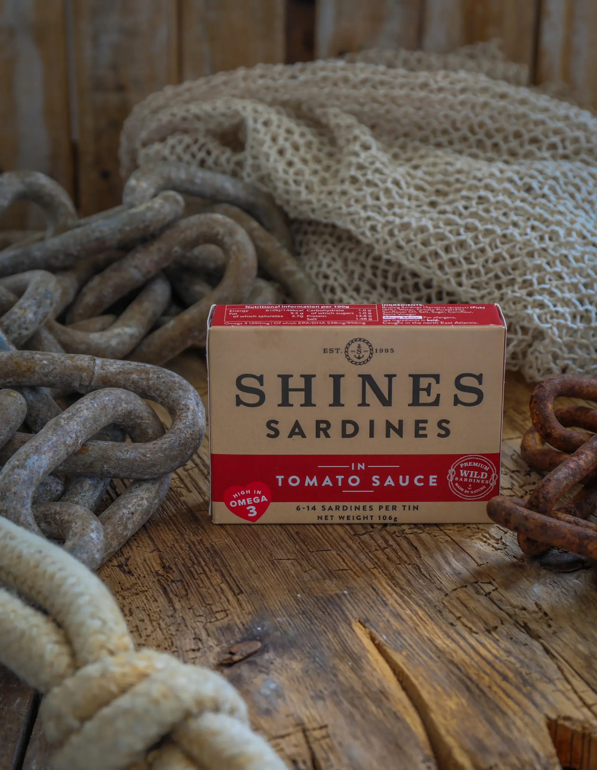 A box of Shines Wild Sardines in Tomato Sauce - 106g rests on a rustic wooden surface, surrounded by thick metal chains, rope, and a fishing net.