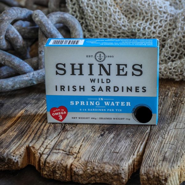 A box of Shines Wild IRISH Sardines in Spring Water (106g) rests on a rustic wooden surface with ropes and fishing net behind it. The packaging notes it's high in Omega 3 and contains 6-14 sardines per tin.