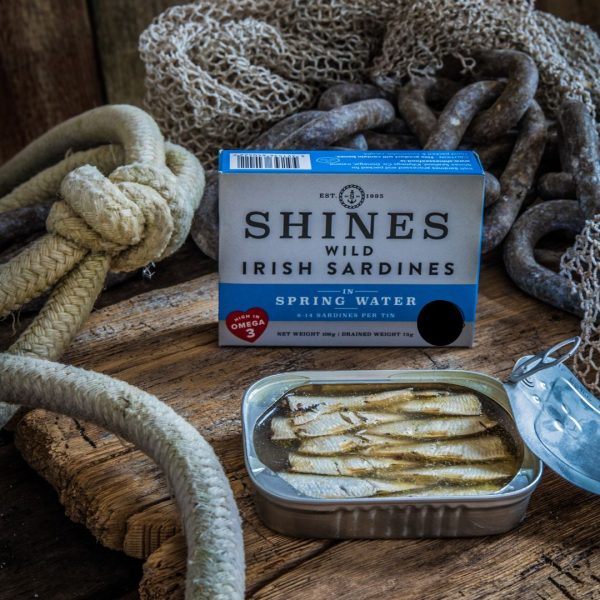 An open tin of Shines Wild IRISH Sardines in Spring Water (106g) rests on a wooden surface beside its box, surrounded by thick ropes and fishing nets for an authentic nautical feel.