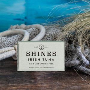 A tin of Shines Wild Irish Tuna in Sunflower Oil -111G sits on a wooden surface, surrounded by fishing net, rope, and dried grass, with a blue sea-like background.
