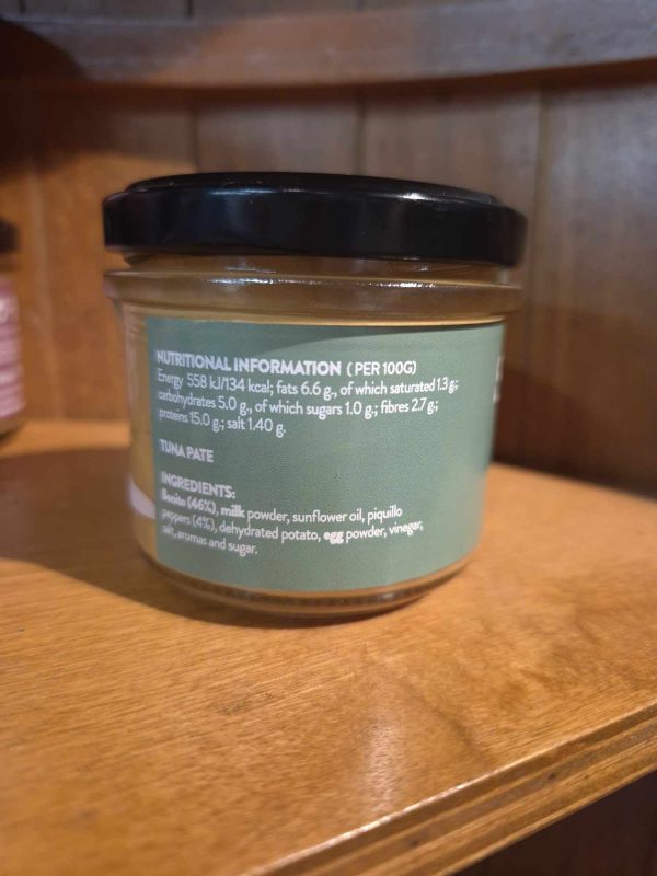 A 100g glass jar of Shines Tuna PÂTÉ with a green label displays nutritional values per 100g and ingredients in white text, resting on a wooden shelf.