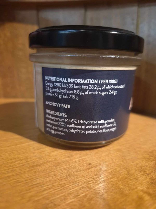 A glass jar with a black lid labelled "Shines Tuna PÂTÉ 100g" sits on a wooden surface. The label displays nutritional information and ingredients, including tuna pâté, sunflower oil, and salt.