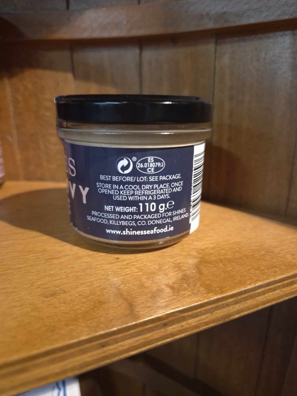 A 100g Shines Tuna PÂTÉ jar with a blue container and black lid sits on a wooden shelf. The label shows storage instructions, a website URL, and a recycling symbol. Only part of the product’s text is visible.