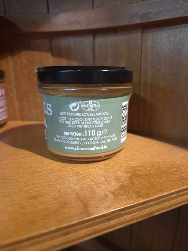 A Shines Tuna PÂTÉ 100g jar with a black lid sits on a wooden shelf, back label facing the camera. The green label displays storage instructions, website, and net weight.