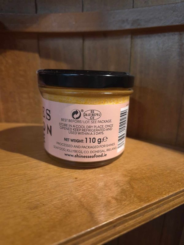 A Shines Tuna PATE 100g jar with a black lid sits on a wooden shelf. Its label shows storage instructions, net weight (100g), and shinessseafood.ie. Some text is only partially visible in the image.
