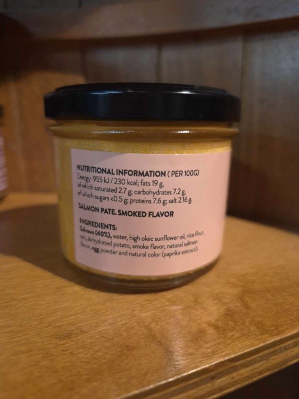 A 100g glass jar of Shines Tuna PÂTÉ with a black lid sits on a wooden shelf. The label lists nutritional info and ingredients: tuna, sunflower oil, rice flour, potato, flavour, lemon, salt, and paprika extract.