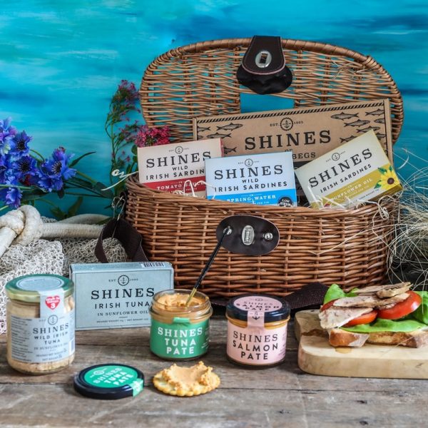 The Howth Hamper, featuring an assortment of Shines fish products like tinned sardines, tuna, pâtés, and jars, is displayed with fish-topped bread and vegetables on a table set against a blue ocean-themed background.