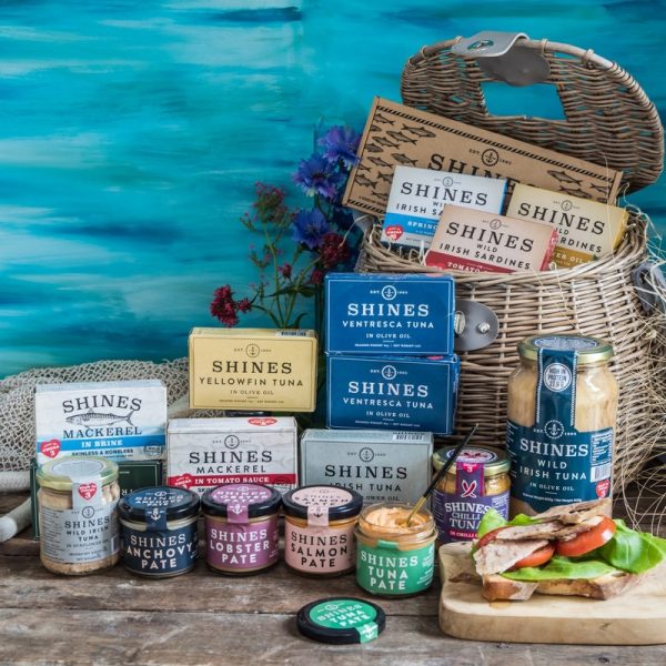 The Killybegs Hamper, featuring tins and jars of Shines tuna, sardines, mackerel, and pâté, is displayed on a table beside a wicker basket, flowers, and an open sandwich against a blue background.