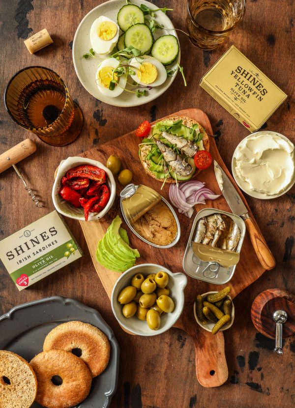 The Killybegs Hamper features a rustic table spread with tinned sardines, avocado slices, red peppers, olives, pickles, cucumber salad, hard-boiled eggs, cream cheese, bagels, and toast topped with sardines.