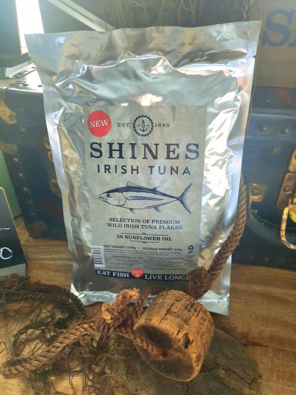 A silver 1kg pouch of Shines Wild Irish Tuna in Sunflower Oil stands upright on a wooden surface beside a fishing net and cork float, with a fish illustration and a red "NEW" badge on the label.