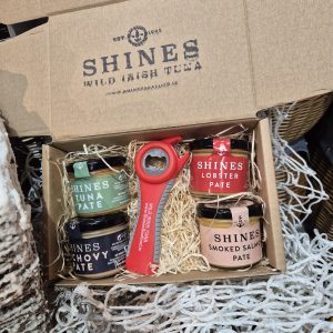 An open Shines Pâté GIFT BOX 4 X100g Jar features four jars of pâté and a red tin opener, all nestled in straw packaging with a fishing net backdrop.