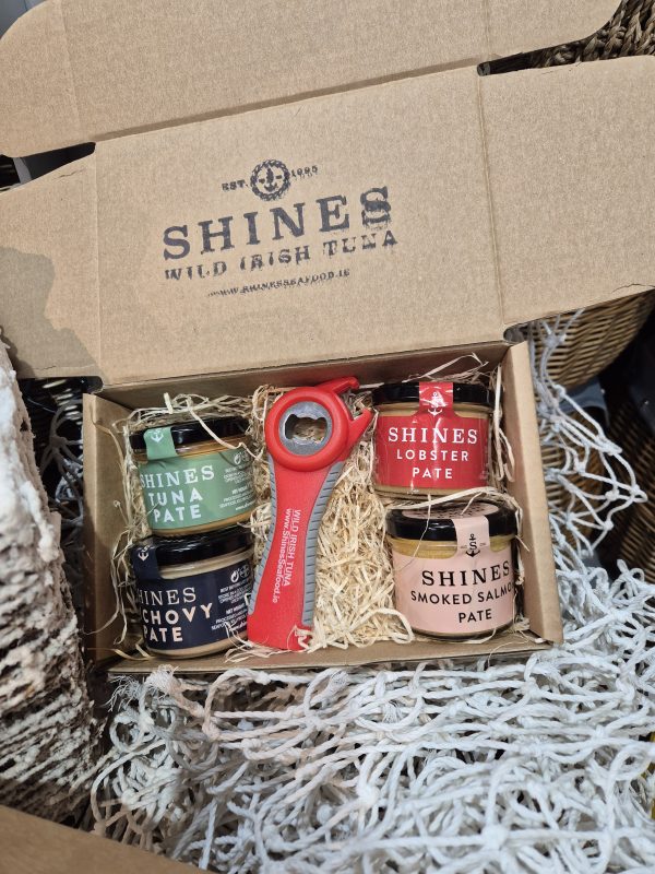 An open Shines Pâté GIFT BOX 4 X100g Jar features four jars of pâté and a red tin opener, all nestled in straw packaging with a fishing net backdrop.