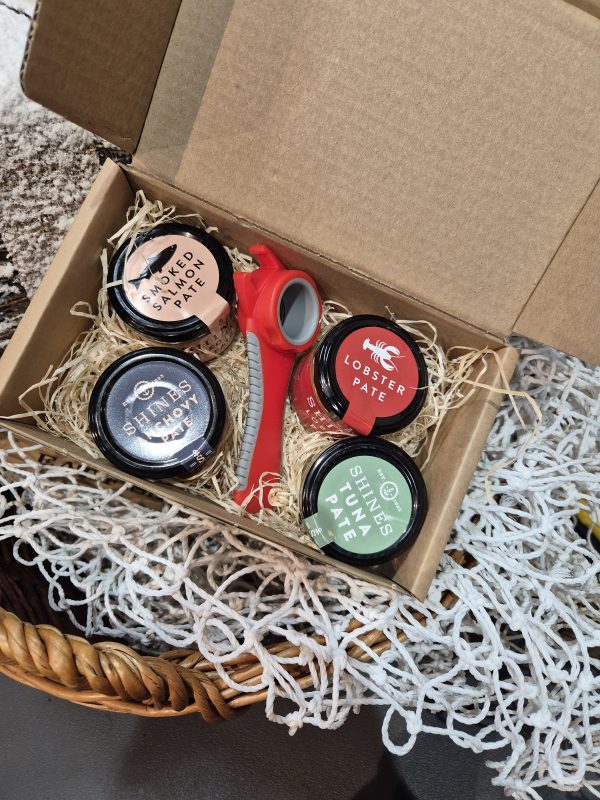 The Shines Pâté Gift Box features four 100g jars of seafood pâté—smoked salmon, lobster, prawn, and tuna—in a woven basket with a red tin opener and decorative straw.