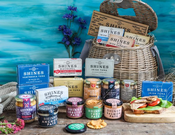A wicker basket filled and surrounded by assorted Shines seafood products, including tins and jars of tuna, mackerel, salmon, and more, with a sandwich and flowers in the background on a blue surface.