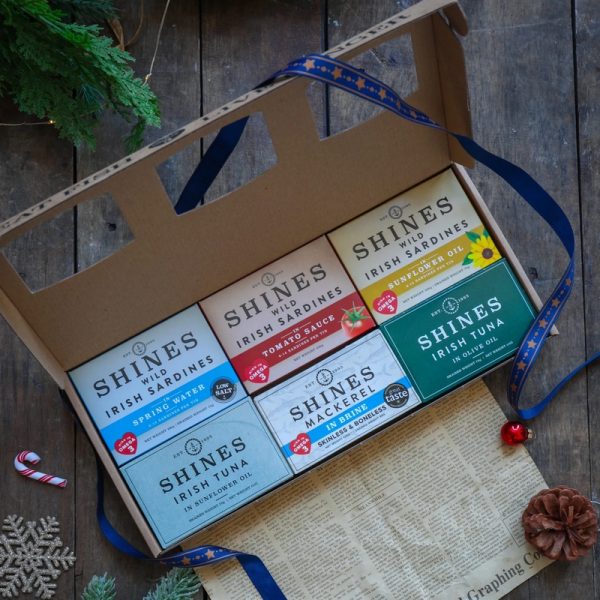 A cardboard gift box containing six tins of Shines tinned fish, including sardines, tuna, and mackerel, is displayed on a wooden table with festive decorations and ribbon nearby.