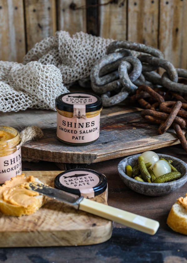 A Shines Pâté GIFT BOX, featuring four 100g jars, sits on a rustic wooden board surrounded by bread, a knife, pickles, onions, and decorative fishing nets and chains in the background.