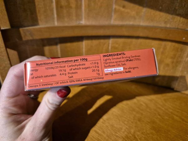 A hand holds a box of Shines LIGHTLY SMOKED IRISH Sardines in Sunflower Oil (106g). The side lists sardines (75%) and sunflower oil as ingredients, nutritional info, and allergy advice for fish.