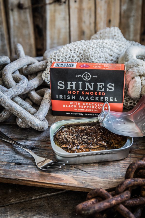 An opened tin of HOT SMOKED Mackerel Fillets with Peppercorns in Sunflower Oil 110g rests by its packaging on a rustic wooden surface, surrounded by fishing nets, metal chains, and a fork.