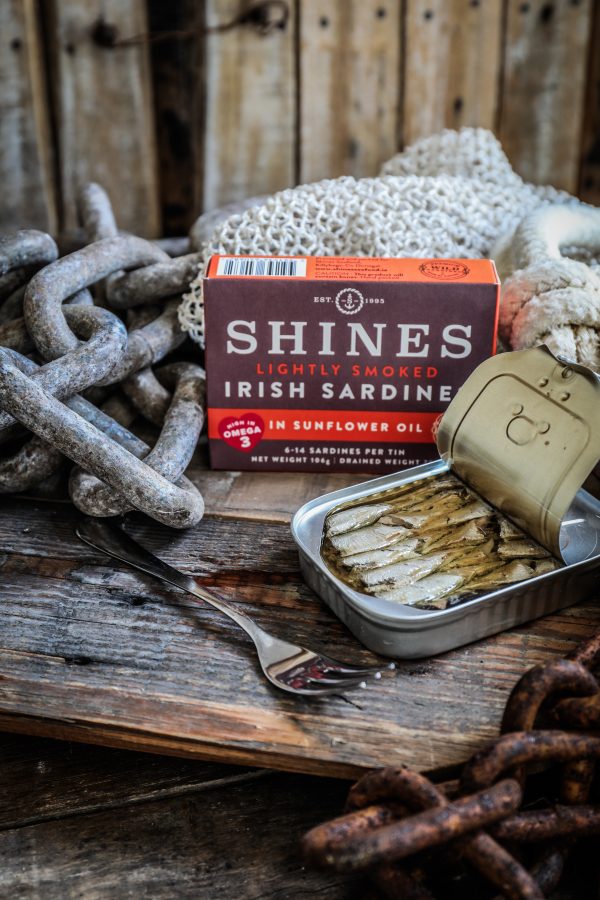 An open tin of HOT SMOKED Mackerel Fillets with Peppercorns in Sunflower Oil 110g rests on a rustic wooden surface beside a fork, with the product box and weathered fishing gear in the background.