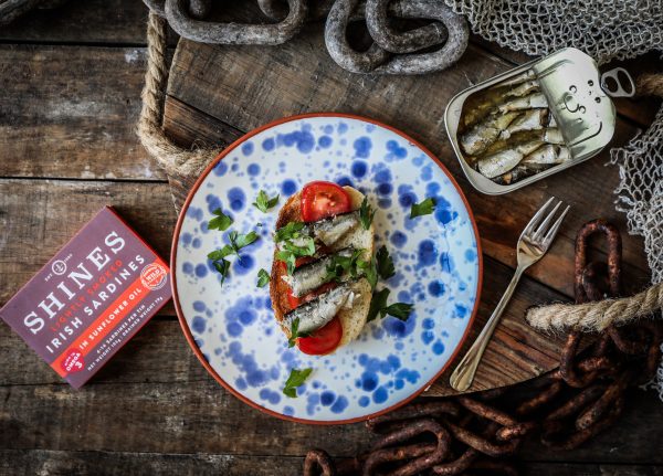 A plate with toast topped with tomato slices, HOT SMOKED Mackerel Fillets with Peppercorns in Sunflower Oil, and herbs sits on a rustic table beside the mackerel tin, a fork, a red box, ropes, and metal chains.