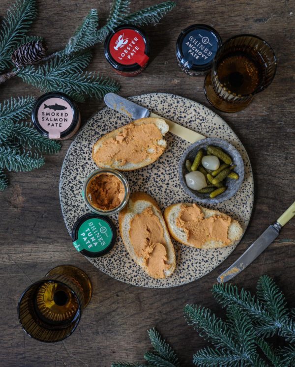 The Perfect Picnic Hamper features seafood pâtés on bread, pickles and onions, lobster and smoked salmon pâté jars, brown glasses, cutlery, and pine branches displayed on a wooden table.