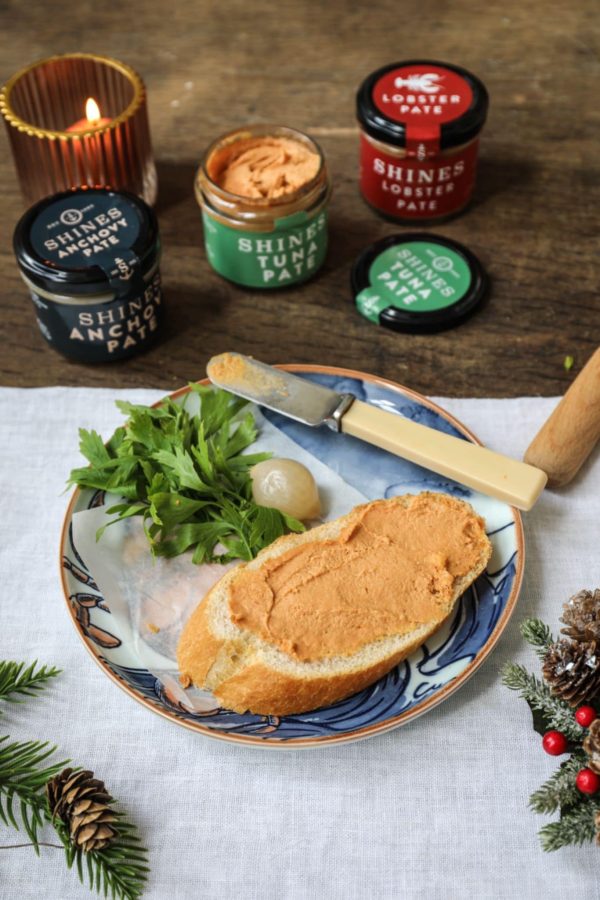 A slice of bread with pâté sits on a plate with parsley and pickled onions, next to jars from the Perfect Picnic Hamper. A lit candle and festive decor adorn the wooden table in the background.