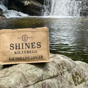A Shines Hessian Bag rests on a rock by a clear river with a waterfall and rocky banks in the background.