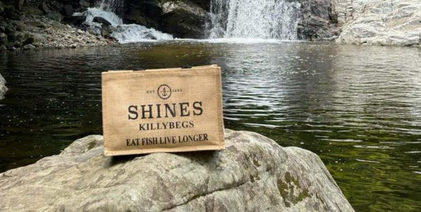 A Shines Hessian Bag rests on a rock by a clear river with a waterfall and rocky banks in the background.