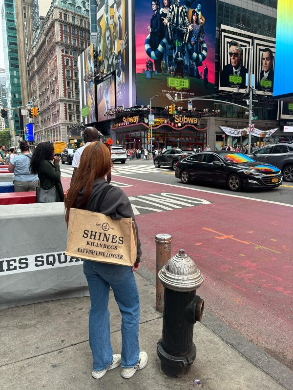 A woman with long hair stands on a New York City pavement, holding the Shines Hessian Bag that says “SHINES KILLVERGS EAT FISH LIVE LONGER,” with Times Square traffic and billboards in the background.