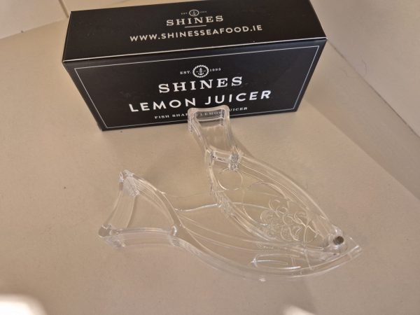 A clear, fish-shaped Shines Lemon Juicer rests on a light surface in front of a black box labelled "Shines Lemon Juicer" with "www.shinesseafood.ie" displayed on the packaging.