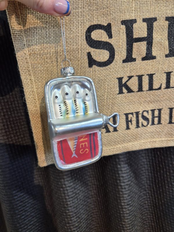 A hand holds the Shines Lemon Juicer, shaped like an open sardine tin with four fish inside, hanging in front of a hessian sign with partially visible black text.