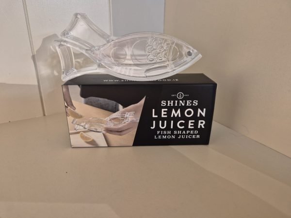 A clear, fish-shaped Shines Lemon Juicer sits on a black and white box labelled "Shines Lemon Juicer," featuring an image of the juicer squeezing lemon slices.