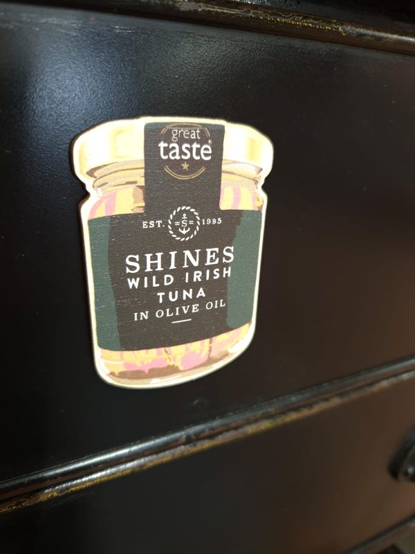 A fridge magnet shaped like a jar on a dark surface, labelled "Shines Tuna" with a "Great Taste" badge at the top.