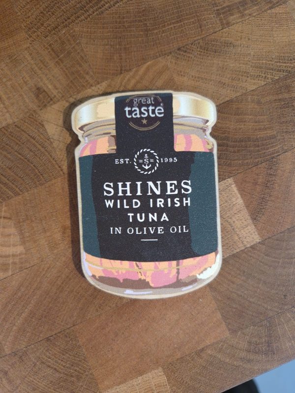 A fridge magnet shaped like a tuna jar labelled "Shines Wild Irish Tuna in Olive Oil" with a "Great Taste" badge, displayed on a wooden surface.