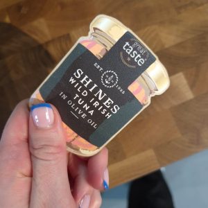 A hand with French-tip blue and pink nails holds a Shines Tuna Jar Fridge Magnet, which features a "great taste" award label, against a wooden background.
