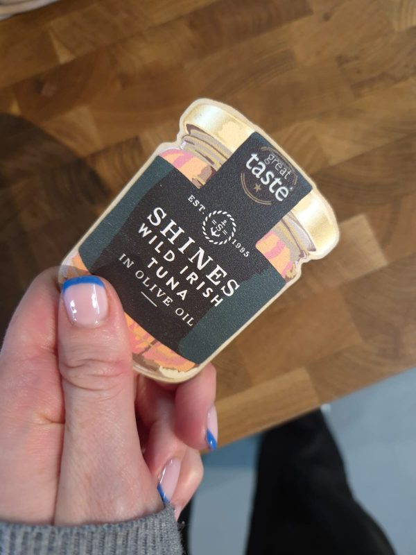 A hand with French-tip blue and pink nails holds a Shines Tuna Jar Fridge Magnet, which features a "great taste" award label, against a wooden background.