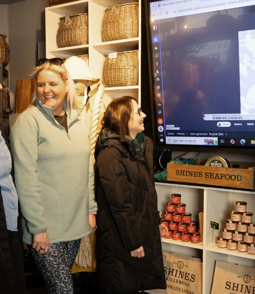 Two women stand smiling beside a large screen in a shop, with stacked baskets and tins labelled "Shines Seafood" on display around them. The atmosphere appears warm and friendly.
