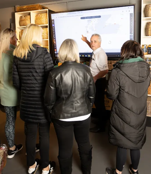 A man in a white shirt points at a large digital map on a screen while four women, dressed in coats, stand facing him and listen. Wicker baskets are displayed on shelves in the background.